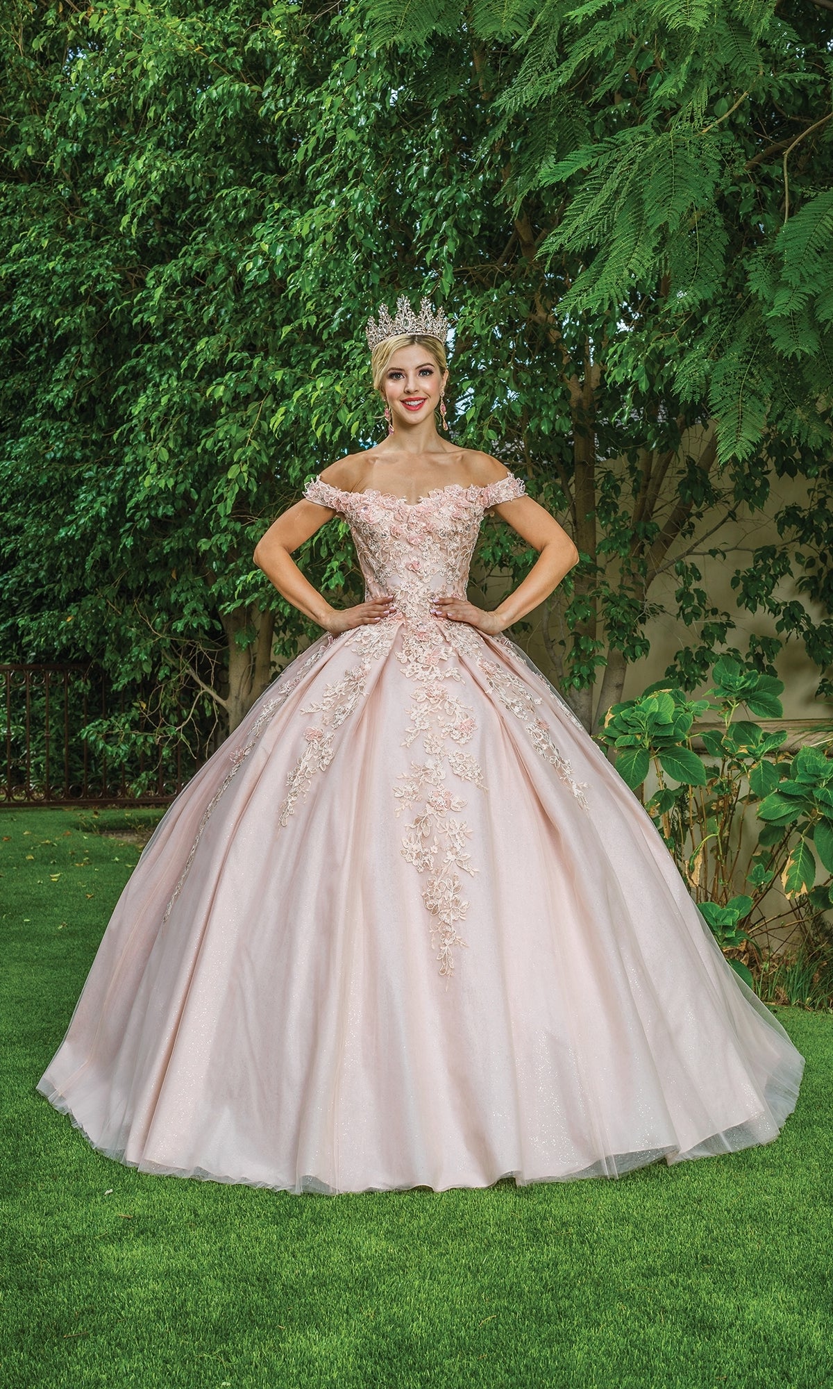 A woman in the Quinceanera Dress 1574 by Dancing Queen, a light pink off-the-shoulder ball gown with floral embroidery and a large crown, stands on grass before green trees.